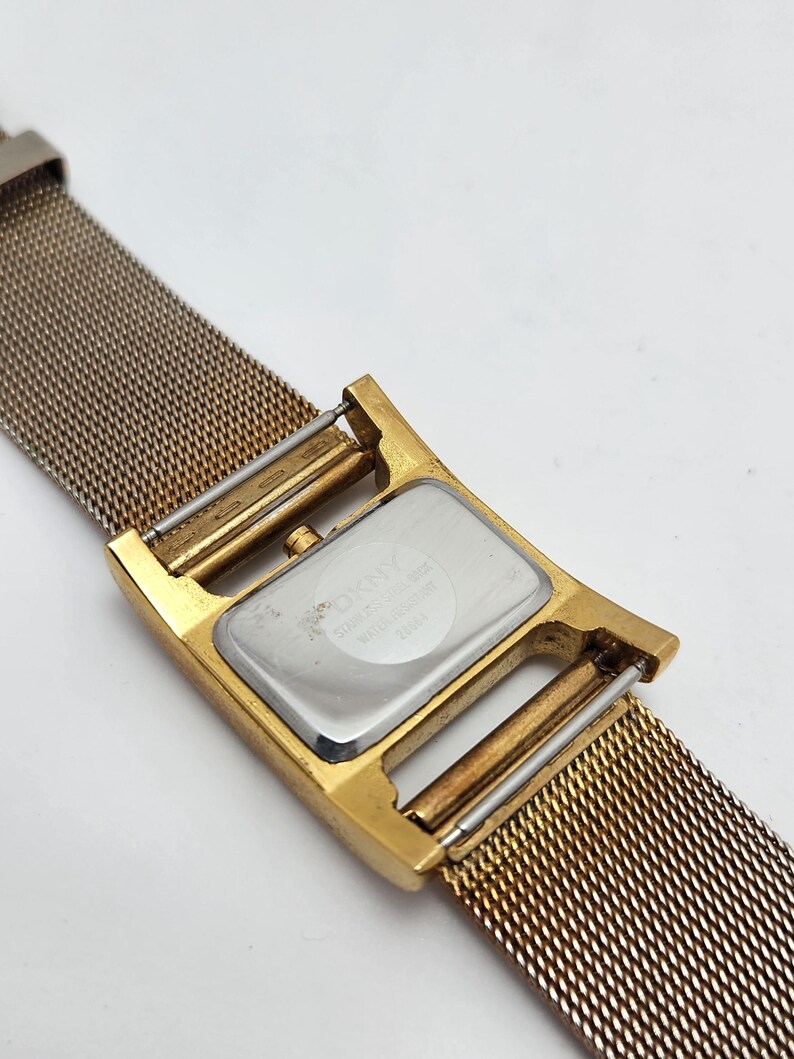 May include: A gold-toned DKNY watch with a mesh band. The watch face is rectangular with a silver backing. The band is a fine mesh of gold-colored metal. The watch is water resistant.
