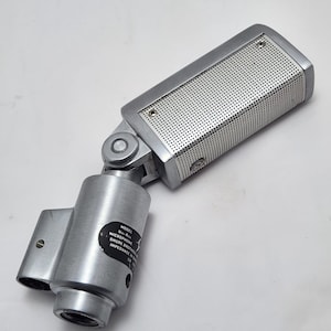 Puede incluir: Micr&oacute;fono plateado vintage con cabezal perforado y base cil&iacute;ndrica. El micr&oacute;fono tiene una junta giratoria y una etiqueta con texto. Probablemente utilizado para grabaci&oacute;n o transmisi&oacute;n.