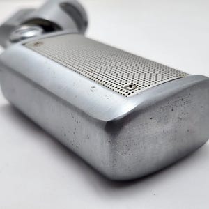 以下が含まれることがあります： 長方形のボディと穴あき金属グリルを備えた、シルバーカラーのヴィンテージマイク。 マイクは丸みを帯びた上部と、使用感のあるブラッシュドメタル仕上げです。
