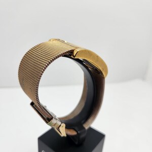 May include: A gold-toned Citizen watch with a mesh band is displayed on a black stand. The watch face is rectangular, and the band is a fine, woven metal. The clasp is visible, and the watch is presented against a white background.