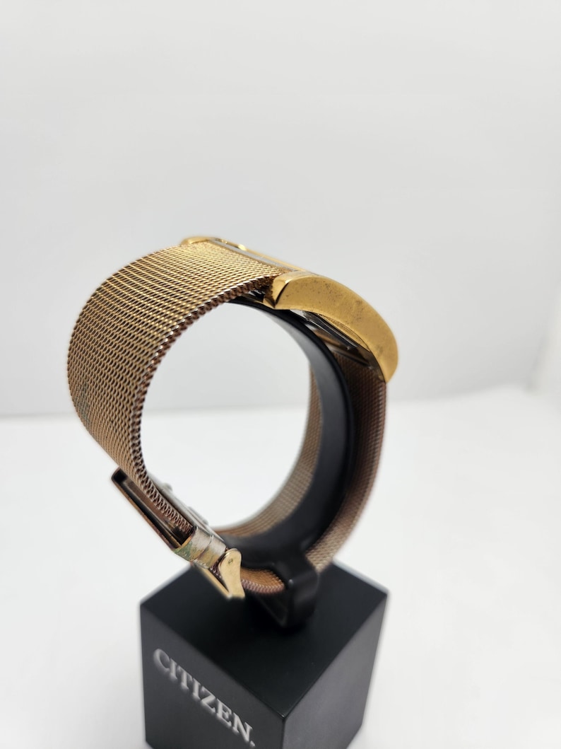 May include: A gold-toned Citizen watch with a mesh band is displayed on a black stand. The watch face is rectangular, and the band is a fine, woven metal. The clasp is visible, and the watch is presented against a white background.
