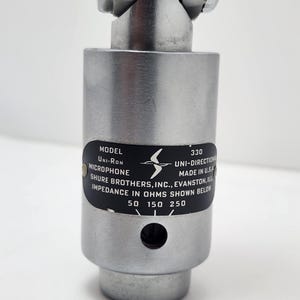 以下が含まれることがあります： 黒いラベルが付いたシルバーカラーのUni-Ronマイク。ラベルには「MODEL UNI-RON MICROPHONE」と「SHURE BROTHERS, INC., EVANSTON, ILL.」と記載されています。マイクには「330 UNI-DIRECTIONAL MADE IN U.S.A.」と記載されています。