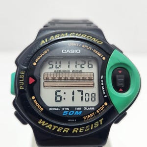 May include: Black Casio digital watch with green and gold accents. The watch face displays the time, date, and various function indicators. Features include alarm, stopwatch, and water resistance. The text "ALARM CHRONO" is visible.