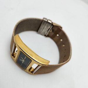 May include: A gold-toned wristwatch with a rectangular black watch face and a mesh band. The watch has a gold-colored bezel with a unique, open design. The band is a woven, metallic material.