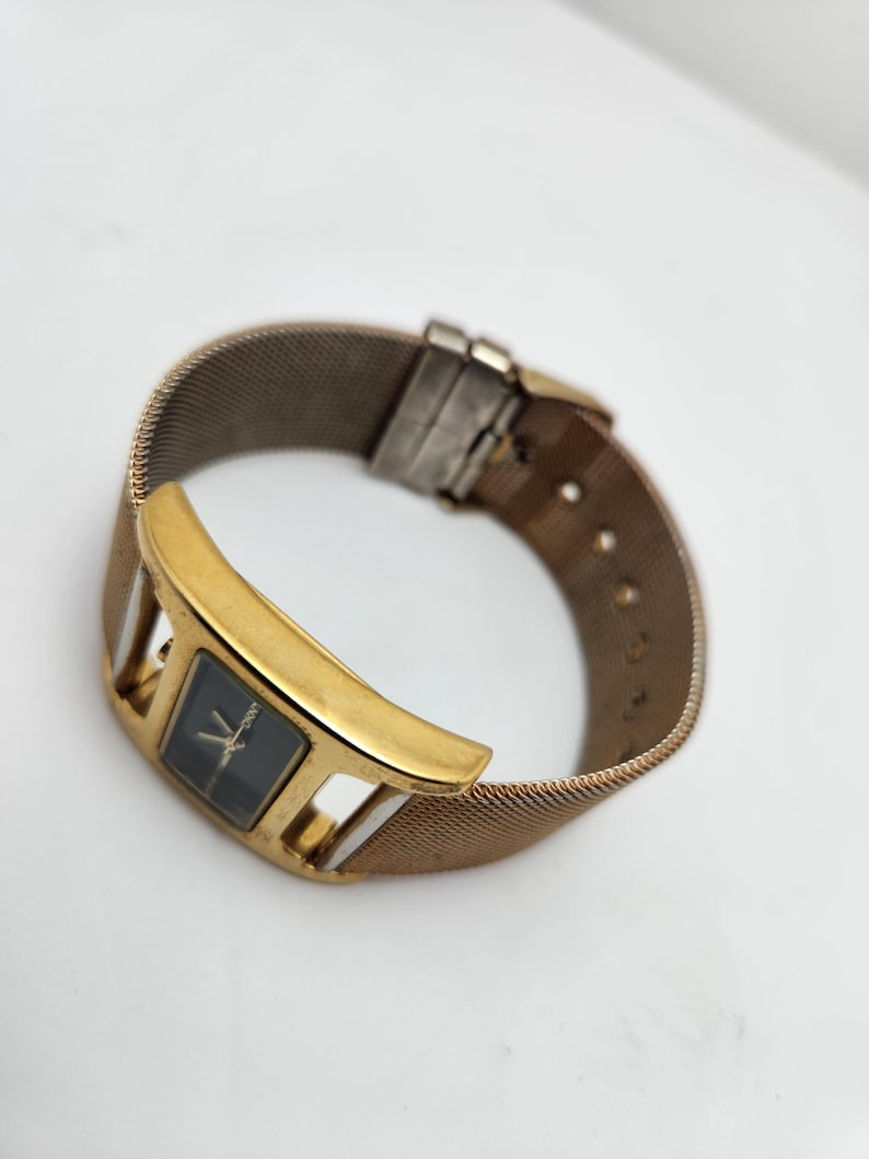 May include: A gold-toned wristwatch with a rectangular black watch face and a mesh band. The watch has a gold-colored bezel with a unique, open design. The band is a woven, metallic material.