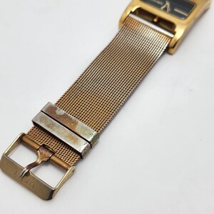 May include: Gold-toned DKNY watch with a rectangular black face and mesh band. The watch features a gold buckle and clasp. The watch band is made of a fine, woven metal.