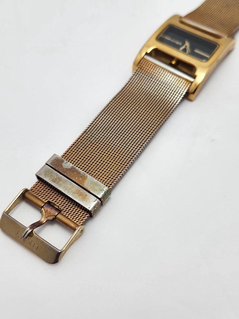 May include: Gold-toned DKNY watch with a rectangular black face and mesh band. The watch features a gold buckle and clasp. The watch band is made of a fine, woven metal.
