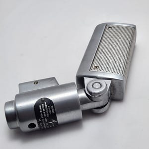 Puede incluir: Un micr&oacute;fono plateado vintage con una cabeza perforada y una junta giratoria. El micr&oacute;fono tiene un cuerpo cil&iacute;ndrico y una etiqueta con texto, incluyendo "UNI-RON" y "SHURE BROTHERS, INC.". Dise&ntilde;ado para grabaci&oacute;n de audio.