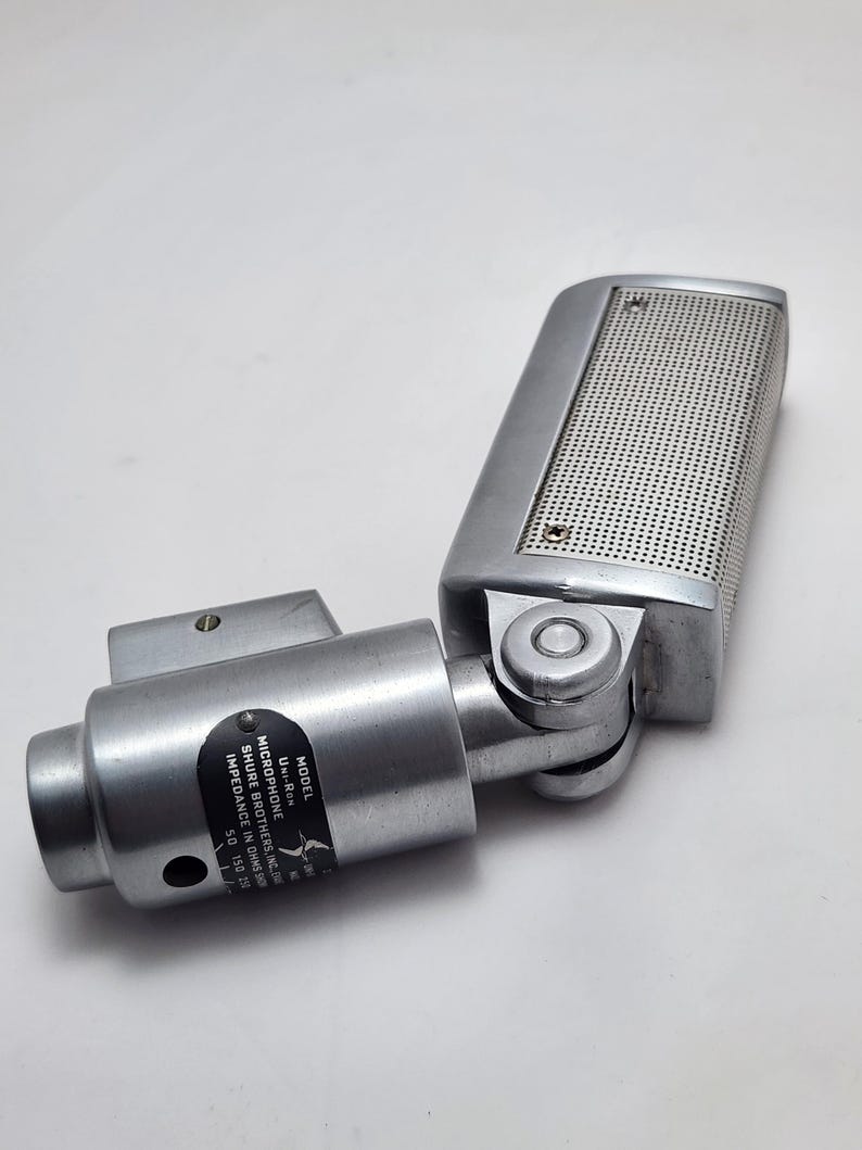 Puede incluir: Un micr&oacute;fono plateado vintage con una cabeza perforada y una junta giratoria. El micr&oacute;fono tiene un cuerpo cil&iacute;ndrico y una etiqueta con texto, incluyendo "UNI-RON" y "SHURE BROTHERS, INC.". Dise&ntilde;ado para grabaci&oacute;n de audio.