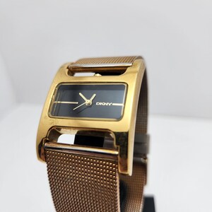 May include: A gold-toned DKNY watch with a rectangular black face and gold hands. The watch features a mesh band and a gold-colored case. The watch is displayed against a white background.