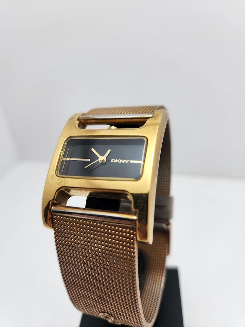 May include: A gold-toned DKNY watch with a rectangular black face and gold hands. The watch features a mesh band and a gold-colored case. The watch is displayed against a white background.