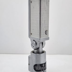Puede incluir: Un micr&oacute;fono plateado vintage con una cabeza rectangular perforada y una base cil&iacute;ndrica. El micr&oacute;fono tiene una junta giratoria y una etiqueta con texto, incluyendo "Shure Brothers Inc."