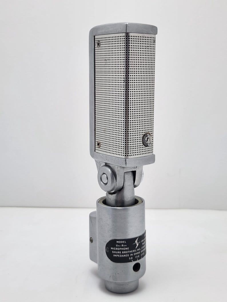 Puede incluir: Un micr&oacute;fono plateado vintage con una cabeza rectangular perforada y una base cil&iacute;ndrica. El micr&oacute;fono tiene una junta giratoria y una etiqueta con texto, incluyendo "Shure Brothers Inc."