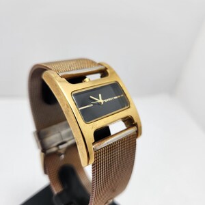 May include: A gold-toned rectangular watch with a black face and gold hands. The watch has a mesh band and is displayed on a black stand. The watch face has the DKNY logo.