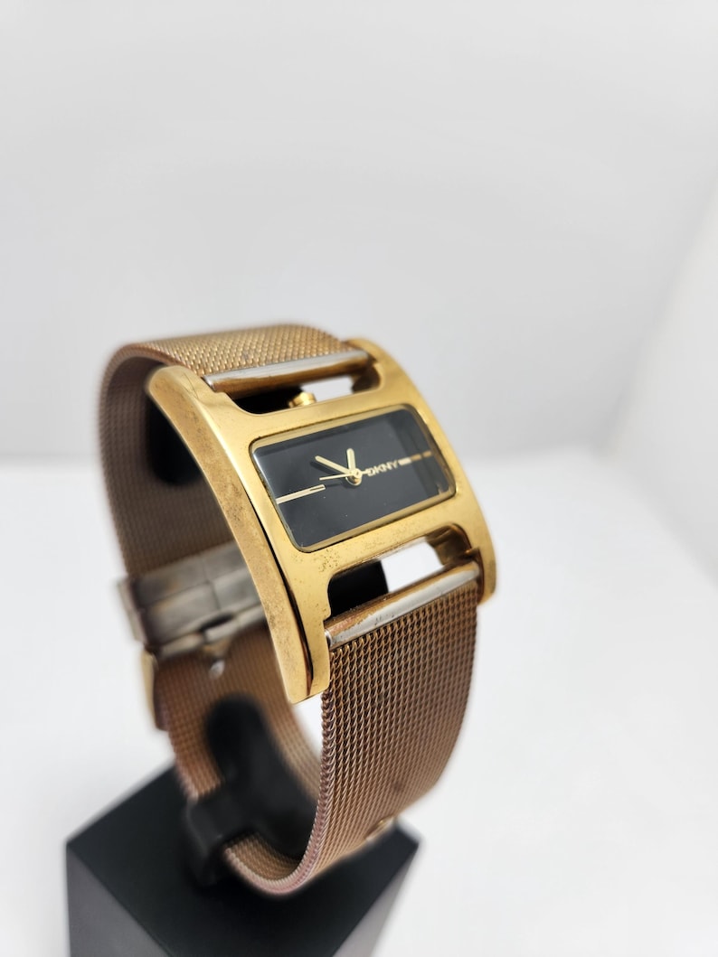 May include: A gold-toned rectangular watch with a black face and gold hands. The watch has a mesh band and is displayed on a black stand. The watch face has the DKNY logo.