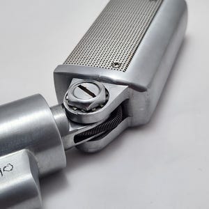 Puede incluir: Un micr&oacute;fono plateado vintage con una cabeza rectangular con una rejilla perforada. El micr&oacute;fono est&aacute; conectado a un cuerpo cil&iacute;ndrico mediante una junta giratoria. El dise&ntilde;o es industrial y retro.