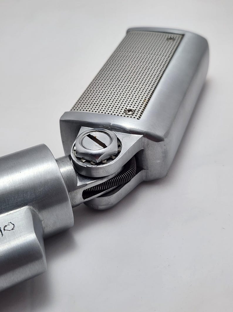 Puede incluir: Un micr&oacute;fono plateado vintage con una cabeza rectangular con una rejilla perforada. El micr&oacute;fono est&aacute; conectado a un cuerpo cil&iacute;ndrico mediante una junta giratoria. El dise&ntilde;o es industrial y retro.