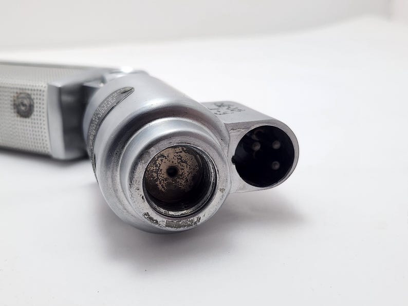 Puede incluir: Primer plano de un micr&oacute;fono plateado con un mango texturizado. El micr&oacute;fono tiene un cuerpo cil&iacute;ndrico con una abertura circular y un conector multipin. El micr&oacute;fono est&aacute; inclinado hacia el espectador.