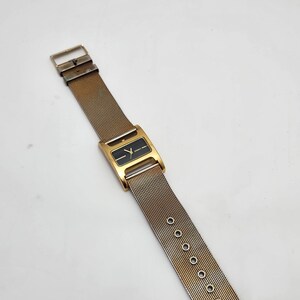 May include: A vintage gold-toned wristwatch with a rectangular black watch face. The watch features a mesh band with a buckle closure. The watch face has a minimalist design with gold-toned hands and markers. The watch is a classic accessory.