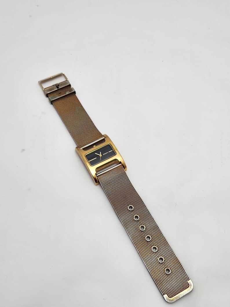 May include: A vintage gold-toned wristwatch with a rectangular black watch face. The watch features a mesh band with a buckle closure. The watch face has a minimalist design with gold-toned hands and markers. The watch is a classic accessory.