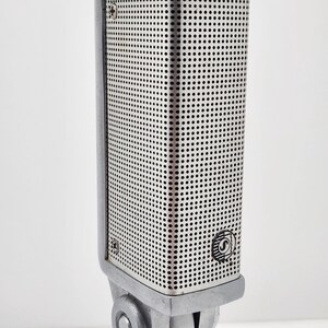 以下が含まれることがあります： 穴あき金属スクリーンを備えたヴィンテージのシルバーマイク。 マイクは長方形のボディで、前面に丸いロゴが付いています。 シルバーのスタンドに取り付けられています。 マイクは録音または放送用に設計されています。