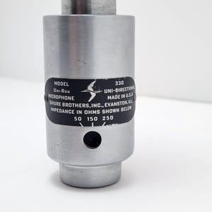 Puede incluir: Un micr&oacute;fono plateado con una etiqueta negra. La etiqueta dice "MODEL Uni-Ron MICROPHONE 330 UNI-DIRECTIONAL MADE IN U.S.A. SHURE BROTHERS, INC., EVANSTON, ILL IMPEDANCE IN OHMS SHOWN BELOW 50 150 250".