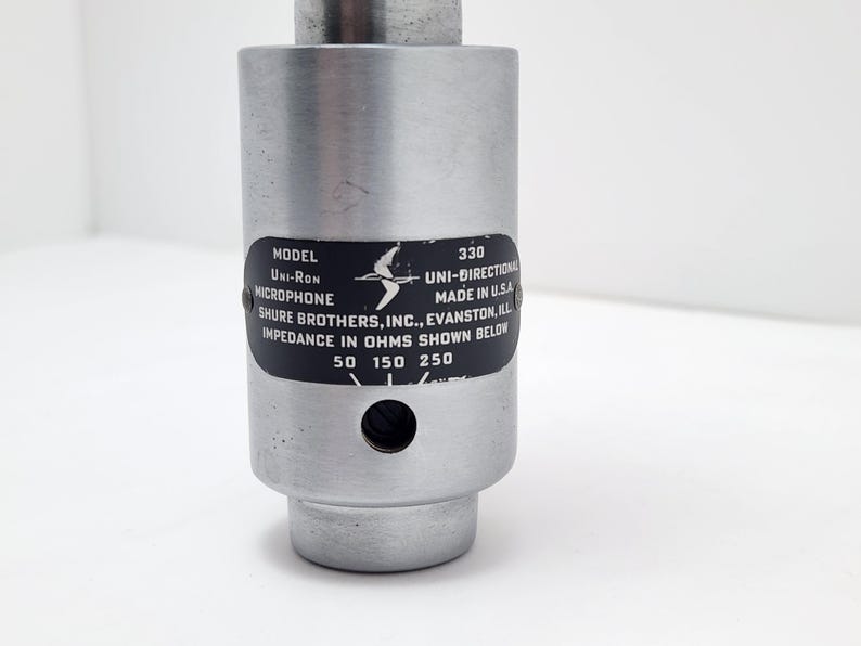 Puede incluir: Un micr&oacute;fono plateado con una etiqueta negra. La etiqueta dice "MODEL Uni-Ron MICROPHONE 330 UNI-DIRECTIONAL MADE IN U.S.A. SHURE BROTHERS, INC., EVANSTON, ILL IMPEDANCE IN OHMS SHOWN BELOW 50 150 250".