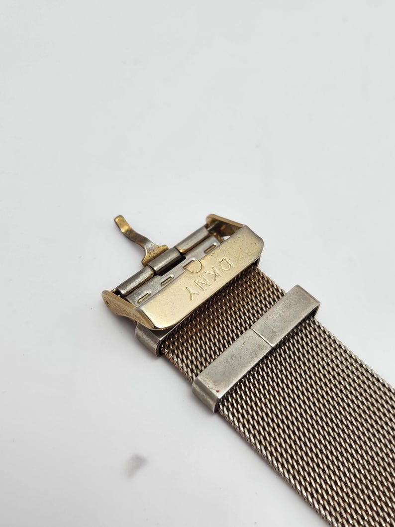 May include: Close-up of a watch band with a mesh design. The band is silver and gold-toned, with a DKNY clasp. The mesh is tightly woven, creating a textured appearance. The clasp is open, showing the inner workings.