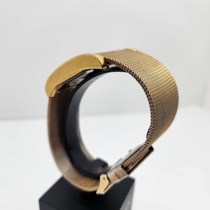 May include: A gold-toned wristwatch with a mesh band and a rectangular face. The watch is displayed on a black stand, showcasing its design and metallic finish. The band features a clasp closure.
