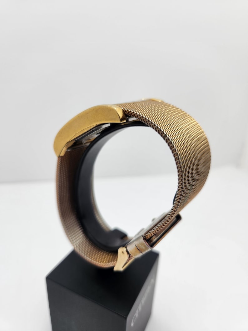 May include: A gold-toned wristwatch with a mesh band and a rectangular face. The watch is displayed on a black stand, showcasing its design and metallic finish. The band features a clasp closure.