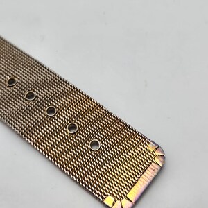 May include: Close-up of a rose gold mesh watch band. The band has a woven texture with several circular holes for adjustment. The edges of the band have a subtle iridescent finish, adding a touch of color.