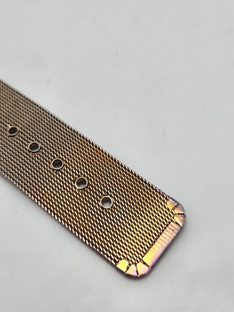 May include: Close-up of a rose gold mesh watch band. The band has a woven texture with several circular holes for adjustment. The edges of the band have a subtle iridescent finish, adding a touch of color.