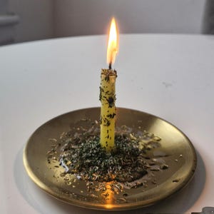 May include: A thin, pale yellow candle with a lit flame, coated in dried green herbs, standing upright in the center of a round, polished brass plate. The base of the candle is surrounded by a cluster of loose, dried green herbs and melted wax.