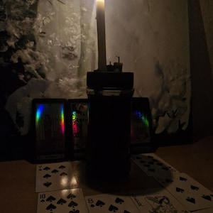 Pode incluir: Uma vela acesa em um suporte preto está em frente a cartas de tarô e cartas de baralho. As cartas estão dispostas sobre uma superfície de madeira, com a chama da vela emitindo um brilho quente. As cartas estão dispostas em uma leitura.
