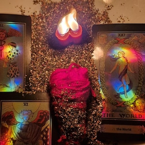 Könnte beinhalten: Eine brennende rote Kerze mit herzförmiger Flamme steht auf getrockneten Kräutern, umgeben von Tarotkarten. Die Karten zeigen farbenfrohe Illustrationen und die Titel "Die Welt" und "Die Liebenden". Die Szene suggeriert ein mystisches oder spirituelles Thema.