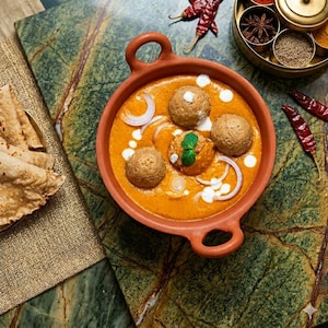 May include: A terracotta bowl filled with orange curry containing three round meatballs, garnished with white onion slices, cream swirls, and a green herb leaf. Beside the bowl are folded flatbreads and dried red chili peppers.