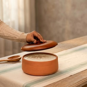 May include: A round terracotta bowl with horizontal ribbed detailing on the exterior, filled with a light-colored substance. A matching circular wooden lid is held slightly above the bowl, and a wooden spoon rests nearby on the surface.