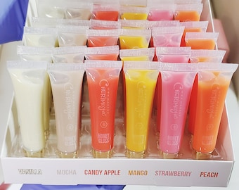 Może przedstawiać: Przezroczyste plastikowe pudełko wypełnione małymi tubkami z błyszczykiem do ust w różnych kolorach, w tym wanilia, mokka, jabłko cukrowe, mango, truskawka i brzoskwinia. Tubki są oznaczone nazwą marki "CHERIMOJa" i smakiem.