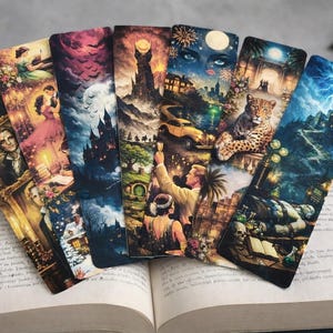 Puede incluir: Un conjunto de marcapáginas de temática fantástica abanicados sobre un libro abierto. Cada marcapáginas presenta una ilustración colorida diferente, incluyendo retratos, escenas románticas y paisajes con castillos.