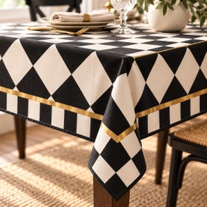 Könnte beinhalten: Eine quadratische Tischdecke mit einem schwarz-weißen Rautenmuster auf der Oberseite, die von einem horizontalen Goldstreifen und einem unteren Abschnitt mit schwarz-weißen Karos gesäumt und über einen Holztisch drapiert ist.