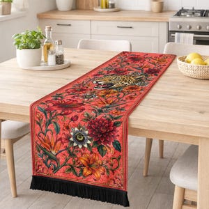 May include: A rectangular table runner featuring a vibrant red background with a detailed print of a snarling leopard head surrounded by various colorful flowers including lilies and passion flowers, finished with black fringe on the ends.