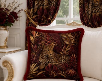 Puede incluir: Un cojín cuadrado con un estampado detallado de un leopardo descansando entre follaje tropical de tonos rojo oscuro y dorado. El cojín está enmarcado por un borde de terciopelo rojo intenso y se encuentra sobre un sofá blanco con cortinas.