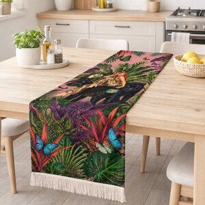 Op de afbeelding: Een rechthoekige tafelloper met een levendige jungleprint met een zwarte panter, een persoon en kleurrijke vlinders. Het ontwerp bevat weelderig groen gebladerte, rode planten en blauwe vlinders op een roze achtergrond, afgewerkt met witte franjes.