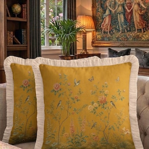 Pode incluir: Duas almofadas amarelo mostarda com padrões florais e de pássaros. As almofadas têm uma borda com franjas creme. Estão em um sofá claro em uma sala com uma tapeçaria e uma estante.