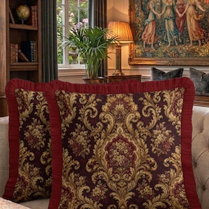 May include: Two square throw cushions featuring a deep burgundy and gold damask pattern with intricate floral motifs. Each cushion is finished with a pleated burgundy fabric ruffle border along the edges, creating a textured frame for the ornate design.