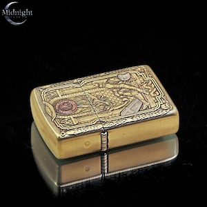 May include: A rectangular, brass-colored lighter with intricate engraved dragon and architectural designs. The lighter has a hinged lid and a reflective surface below. The top left corner has the text "Midnight" above "store co."
