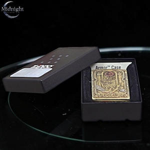 May include: A gold-colored Zippo lighter with an intricate design, displayed in a black box. The lighter features a detailed emblem and the words "Armor Case". The box is open, revealing the lighter against a black interior.