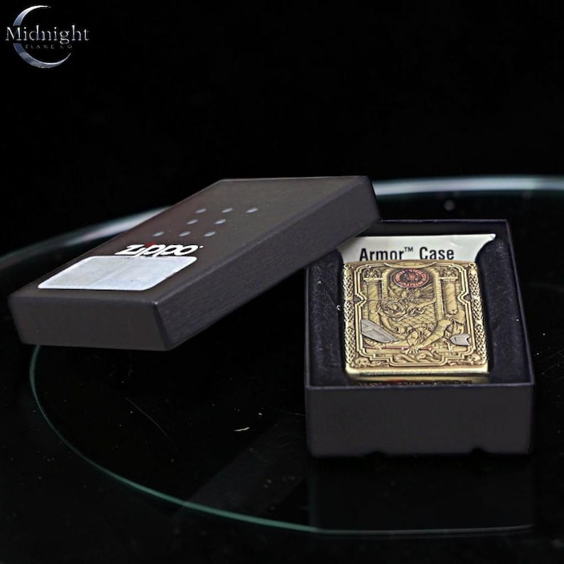 May include: A gold-colored Zippo lighter with an intricate design, displayed in a black box. The lighter features a detailed emblem and the words "Armor Case". The box is open, revealing the lighter against a black interior.
