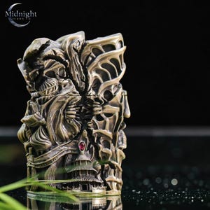 K&ouml;nnte beinhalten: Ein aufwendig geschnitztes Feuerzeug in Bronzefarbe mit detailliertem Reliefdesign. Das Feuerzeug zeigt eine Struktur im Pagodenstil, Figuren und einen roten Edelstein. Die Oberseite des Feuerzeugs hat ein Design, das einem Fl&uuml;gel &auml;hnelt. Das Logo "Midnight Trade Co" ist sichtbar.