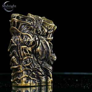 K&ouml;nnte beinhalten: Ein detailliertes, bronzefarbenes Feuerzeug mit einem komplizierten, strukturierten Design. Das Feuerzeug hat ein komplexes, skulpturales Aussehen mit dunklem Hintergrund. In der oberen linken Ecke steht der Text "Midnight Tide Co."